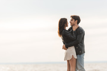 Young man hugging brunette girlfriend in leather jacket with sky and sea at background