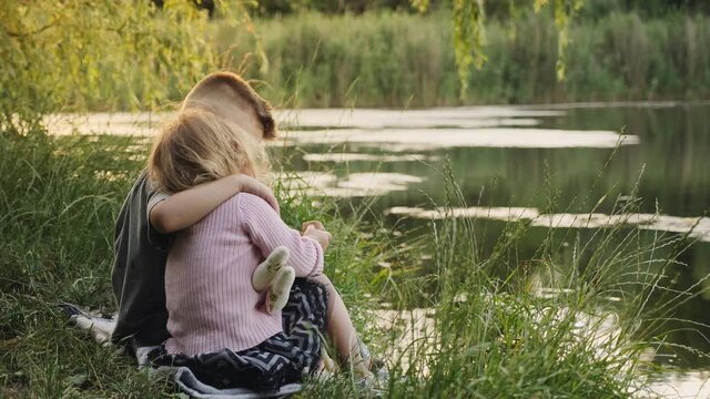 A little boy hugs his little sister while sitting on the shore of a small lake. Happy childhood, carefree pastime