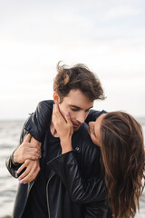 Young woman touching cheek and embracing boyfriend in leather jacket near sea