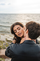 Fototapeta na wymiar Man in black leather jacket kissing girlfriend in neck near sea on beach