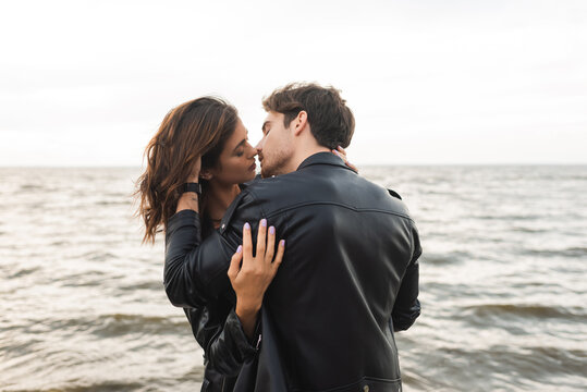 Man in leather jacket kissing girlfriend with sea at background