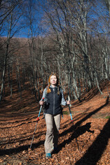 A girl in hiking clothes with trekking poles walks in the mountains in the beech forest.