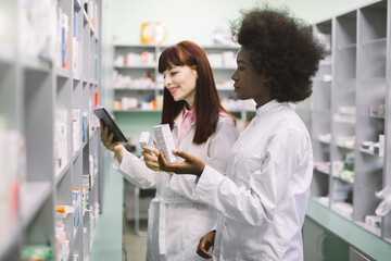 Obraz premium Medicine, pharmaceutics, health care and people concept. Two young multiethnic female pharmacist, African and Caucasian, taking medications from the shelf, using tablet in pharmacy