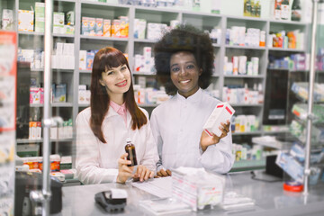 Portrait of two smiling friendly multiethnic female pharmacists working in modern farmacy, posing at the counter. African woman holds white box and Caucasian woman holds bottle of syrup
