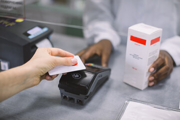 Cropped image of hands of African woman pharmacist and Caucasian woman paying for syrup medicines with credit card in pharmacy, holding credit card. Drug dispensing at pharmacy