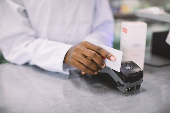 Buying with credit card in the pharmacy. Cropped image of hands of African woman pharmacist swiping credit card of client and a terminal, making payment for drugs
