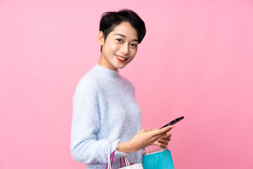 Young Asian girl over isolated pink background holding shopping bags and writing a message with her cell phone to a friend