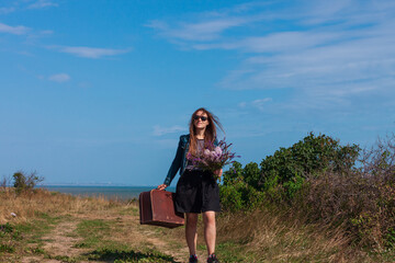 Young woman with long brown hair in denim jacket, black skirt, vintage suitcase,purple wild flowers bouquet off-road sea landscape. Hitchhiker on countryside trip. Lifestyle photo lady walking outdoor