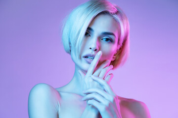 Blond girl model with stylish trendy haircut  looking at camera and touching her lips in neon light at purple studio background.
