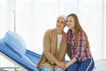 Young beautiful daughter sit beside mother who fight to the cancer for support her on hospital bed in clean and clear ward room, Breast cancer awareness month campaign concept with pink ribbon.