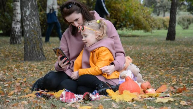 Mom with a Small Daughter Looking Photo on the Phone in the autumn city Park, family love concept