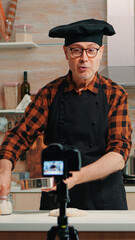 Cheerful elderly baker man filming cooking vlog in home kitchen. Retired blogger chef influencer using internet technology communicating, shooting blogging on social media with digital equipment