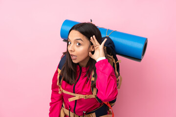 Young mountaineer girl with a big backpack over isolated pink background listening to something by putting hand on the ear