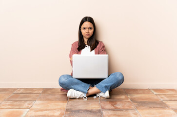 Young mixed race woman with a laptop sitting on the floor with unhappy expression