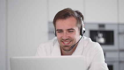Young man wearing headset speaking and watching business webinar sitting in kitchen
