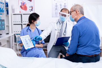 Patient looking at radiography of his skeleton during consultation with doctor in hospital wearing protection against covid-19. Medical examination for infections, disease and diagnosis.