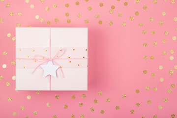 Top view of pastel pink gift box with bow and star on coral background decorated with golden confetti. Flat lay . Holiday festive concept. Copy space.