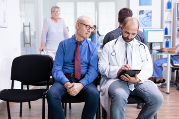 Doctor explaining test results to senior man in hospital waiting area. Disabled senior woman with walking frame arriving for medical examination.