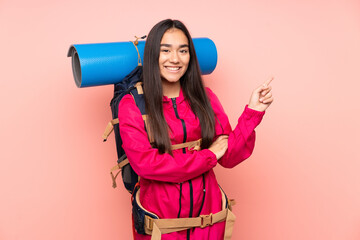 Young mountaineer Indian girl with a big backpack isolated on pink background pointing finger to the side