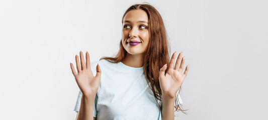 Fototapeta na wymiar Stop. The woman raises her hands to the camera. The model feels confused and does not know what to choose. Beautiful sweet brunette girl with fashion makeup purple lips on an isolated gray background