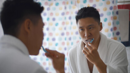 Asian young man with toothbrush cleaning teeth and looking mirror in bathroom.