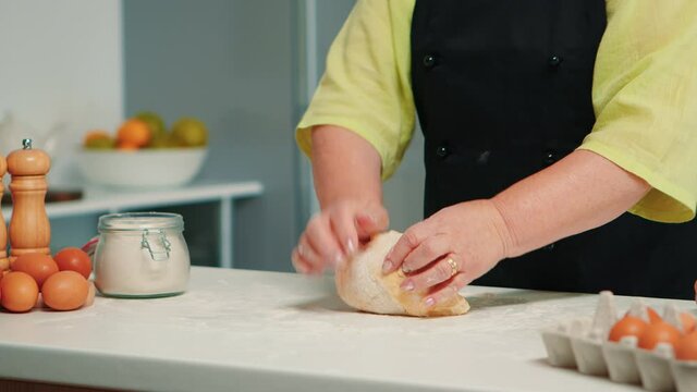 Grandmother hands preparing homemade cookies in modern kitchen kneading on the table. Retired elderly baker with bonete mixing ingredients with wheat flour for baking traditional cake and bread