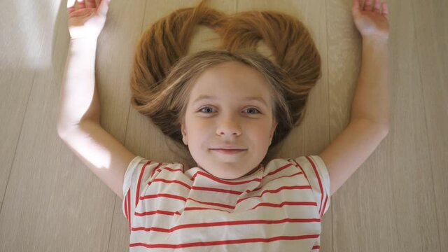 Little funny kid lying on the floor and looking into camera at room. Happy smiling blonde girl relaxing at home. Close up positive emotions of cute child on her face. Top view Slow motion