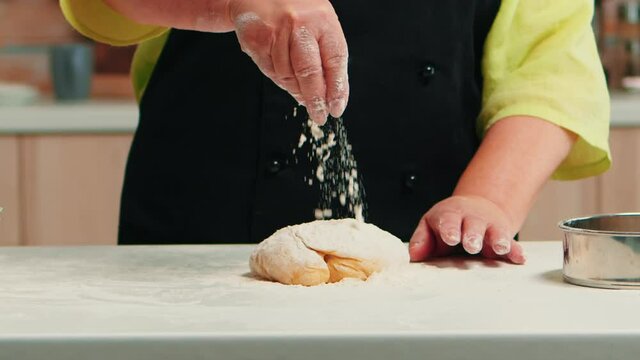 Process of preparing the dough for homemade bread. Close up of retired senior chef with bonete and uniform sprinkling, sieving sifting wheat flour with hand baking homemade pizza and cakes.