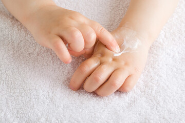 Baby girl finger applying moisturizing cream on hand on white towel. Care about children clean and soft body skin. Front view. Closeup.