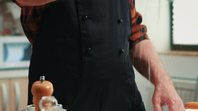 Baker adding flour on the pile by hand preparing bread dough. Close up of retired elderly chef with bonete and uniform sprinkling, sieving spreading rew ingredients baking homemade pizza and cackes.