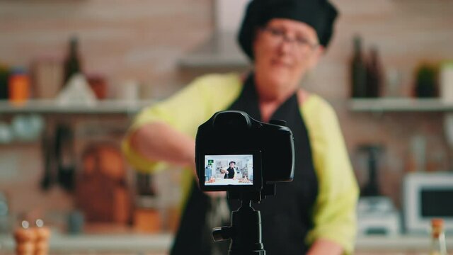 Old lady baker presenting food recipe on cooking podcast from kitchen. Retired blogger chef influencer using internet technology communicating, shooting blogging on social media with digital equipment