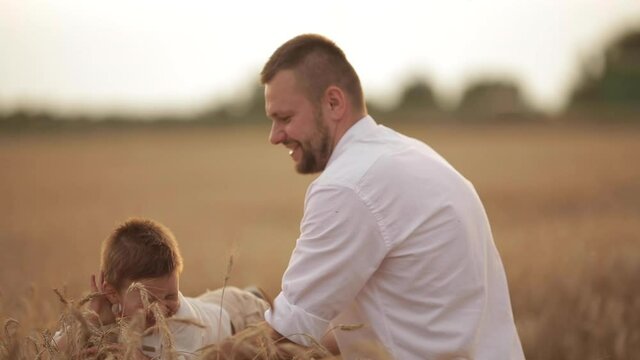 4K stock video of a loving father in white shirt playing with his laughing little son on his arms above spikelets of wheat. Enjoying summer evening in the wheat field together. Family values.