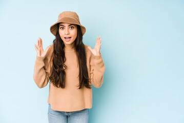 Obraz na płótnie Canvas Young indian woman wearing a hat isolated on blue background receiving a pleasant surprise, excited and raising hands.