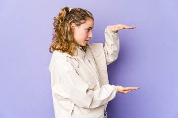 Young caucasian woman on purple background shocked and amazed holding a copy space between hands.