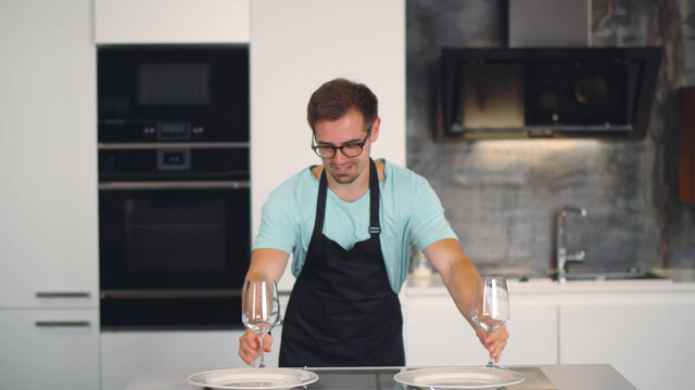 Young handsome man in apron dancing with wine glasses setting table for romantic dinner - Powered by Adobe