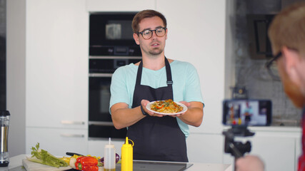 Young man holding plate with pasta and telling how to cook vegetarian dish on camera in kitchen