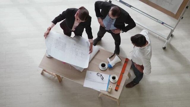 Top view of a team of engineers standing around their desks with paperwork and working equipment. They are discussing construction plan projects in the meeting room.
