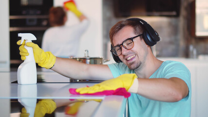 Cheerful man in headphones wiping cooking top with rag and wife cleaning furniture on background
