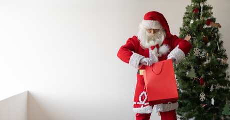 Santa Claus stands near his back near the Christmas tree. Holds a shopping bag in his hands.