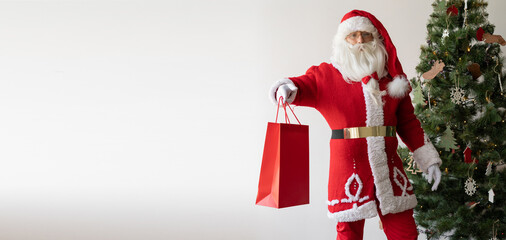 Santa Claus stands near the Christmas tree holding a large shopping bag. Baner.