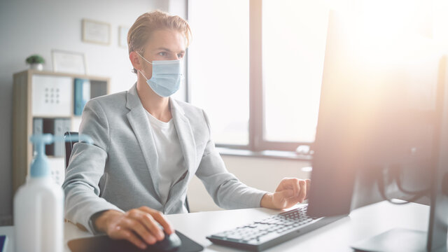 Handsome Stylish Young Man is Wearing a Protective Face Mask and Working at His Desk on a Computer in Office. Blue Eyed Blond Haired Young Male Follows Health and Safety Instructions