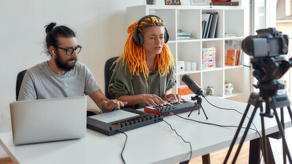 Woman with dreadlocks singing and playing. Female and male blogger making music using synthesizer, drum pad machine and laptop, recording video blog or vlog at home