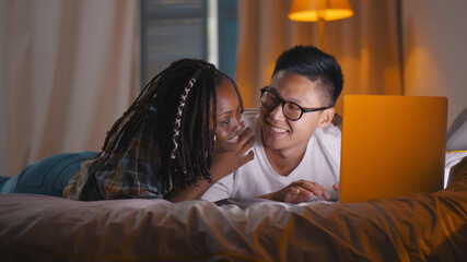 Happy multiethnic couple lying on bed while using laptop computer