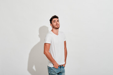 Young man wearing jeans and a white tshit in front of a white wall while he is looking at camera.