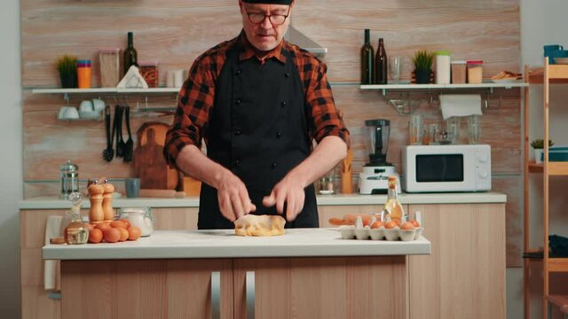 Old man with kitchen apron cooking bread in modern kitchen at home. Retired elderly baker with bonete mixing ingredients with sifted wheat flour kneading for baking traditional cake and bread