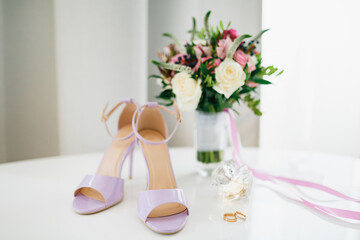 Lilac bride's sandals on the table with the bride and groom's wedding rings and a bouquet of flowers in a vase.