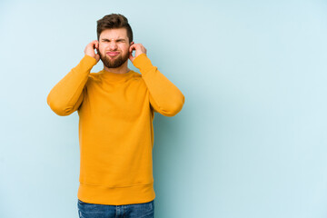 Young caucasian man isolated on blue background covering ears with fingers, stressed and desperate by a loudly ambient.