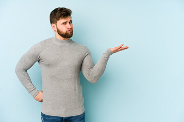 Young caucasian man isolated on blue background doubting and shrugging shoulders in questioning...