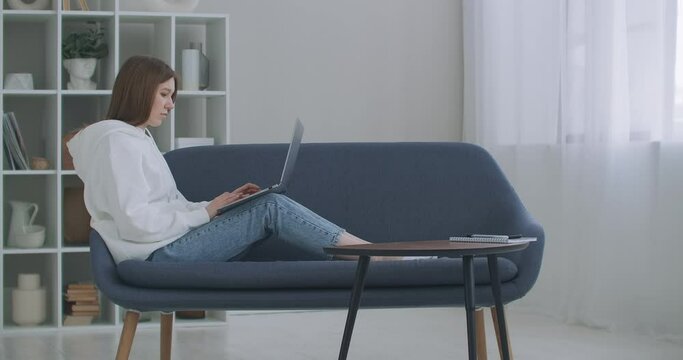 Woman using laptop on couch at home. Thoughtful young woman sitting with computer on couch, looking outside, concentrate on work, feeling bored, need additional motivation, working remotely at home.
