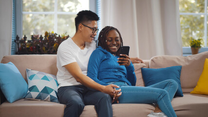 Diverse beautiful couple hugging and using mobile phone while sitting on sofa at home